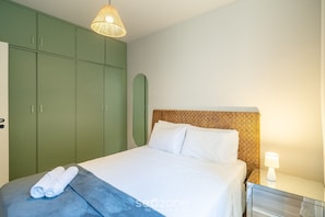 A neatly made bed with white linens, a blue blanket, and rolled towels on a grey bedside table. A bedside lamp is on, and a woven headboard is visible. Green wardrobes are in the background. - Apto Central a 600m da Beira-Mar Norte SGS0106 (Florianópolis)