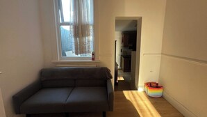 A narrow hallway with a grey sofa, a window with sheer curtains, and a rainbow-colored storage bin.