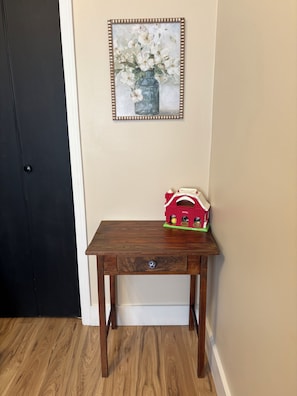 Een houten tafel met een lade, een klein speelboerderijtje erop, en een lijst met een schilderij aan de muur.