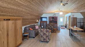 A cozy living room with a wooden ceiling, a sofa, a patterned armchair, a small table, and a kitchen area with a refrigerator.