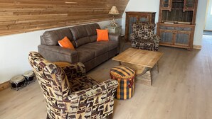 Un salón acogedor con techo de madera, un sofá marrón, un sillón estampado y una pequeña mesa de centro de madera.