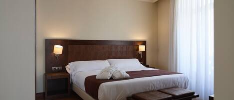 A hotel room with a large bed, two bedside tables, a bench, and a window with sheer curtains.