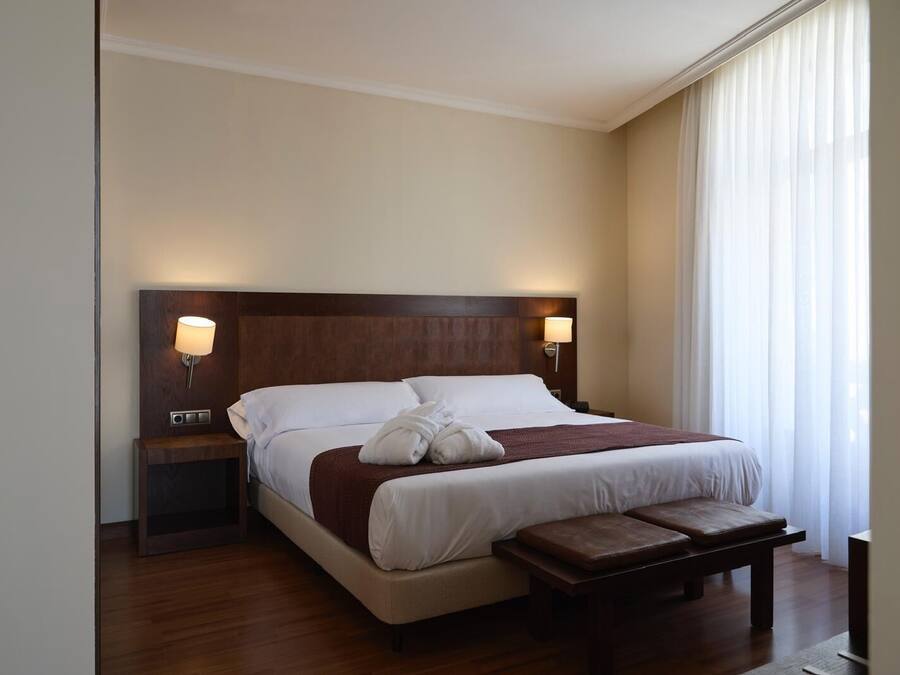A hotel room with a large bed, two bedside tables, a bench, and a window with sheer curtains.