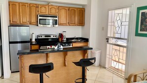 Una cocina con gabinetes de madera, un refrigerador de acero inoxidable, un microondas, una estufa y un lavabo. Hay dos taburetes negros y un bote de basura.
