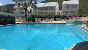 Una piscina azul cristalina con un solárium empedrado, rodeada por un edificio blanco con balcones y una exuberante ladera verde al fondo.