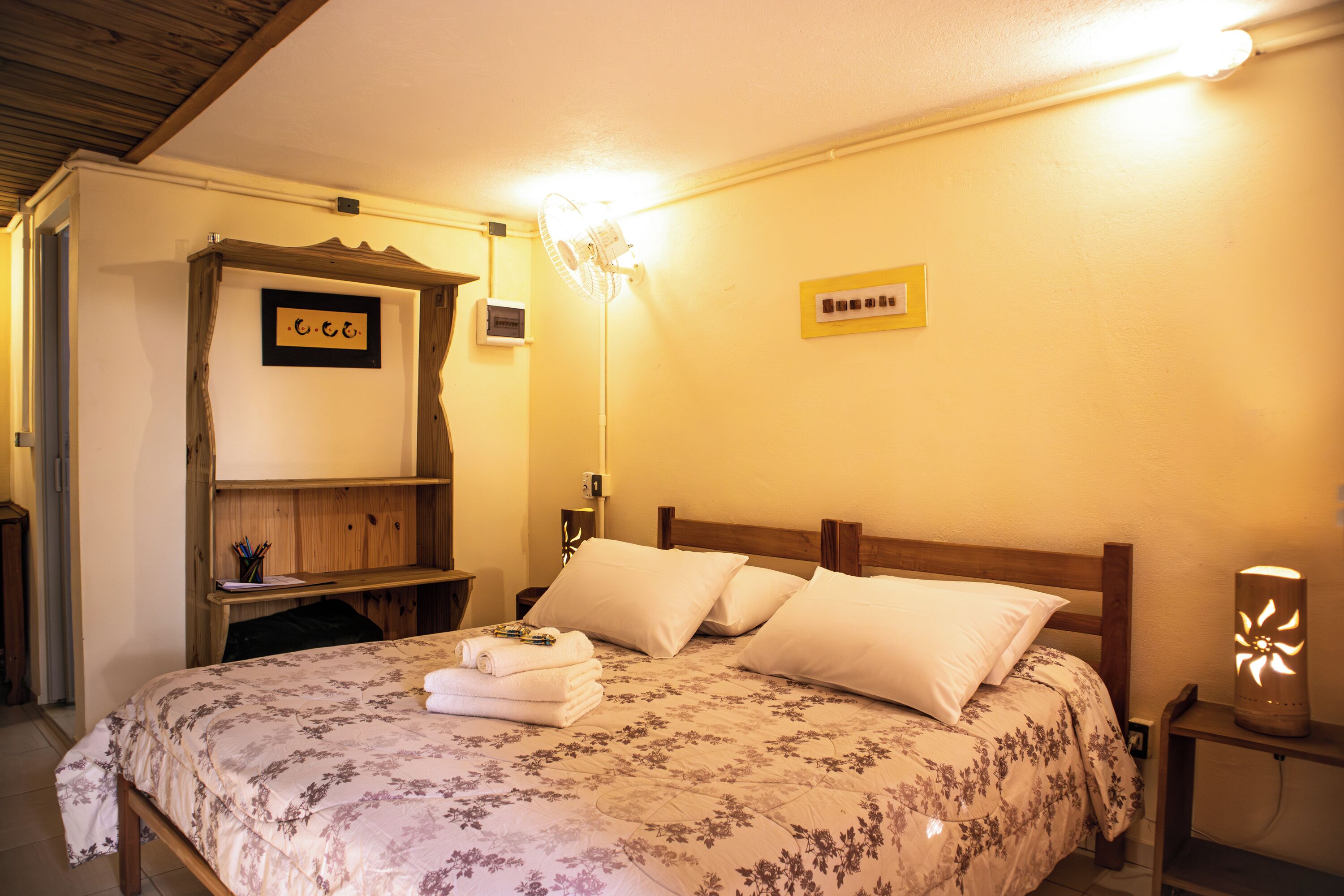 A neatly made bed with a floral bedspread and pillows, a bedside table with a lamp, and a wall-mounted shelf with a small picture and a framed item.