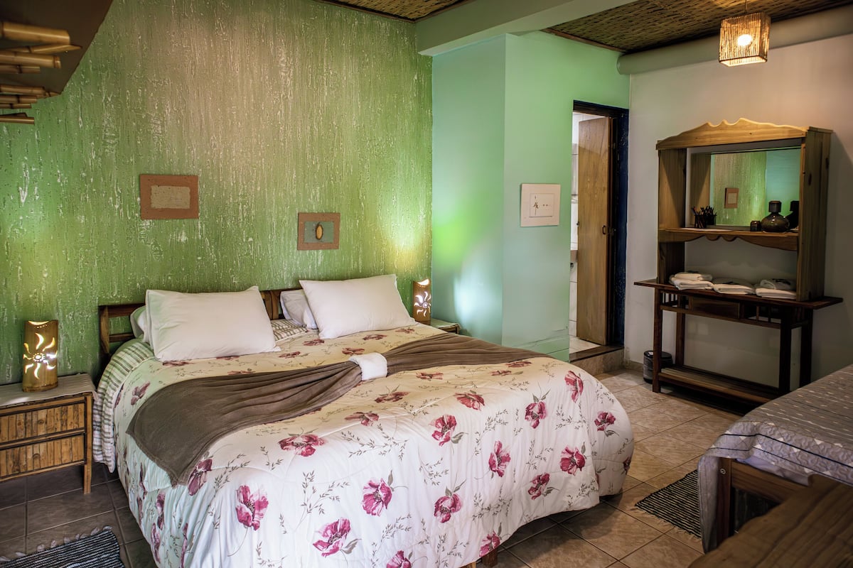 A bedroom with a large bed, a wooden nightstand, a mirror, and a shelf with toiletries.