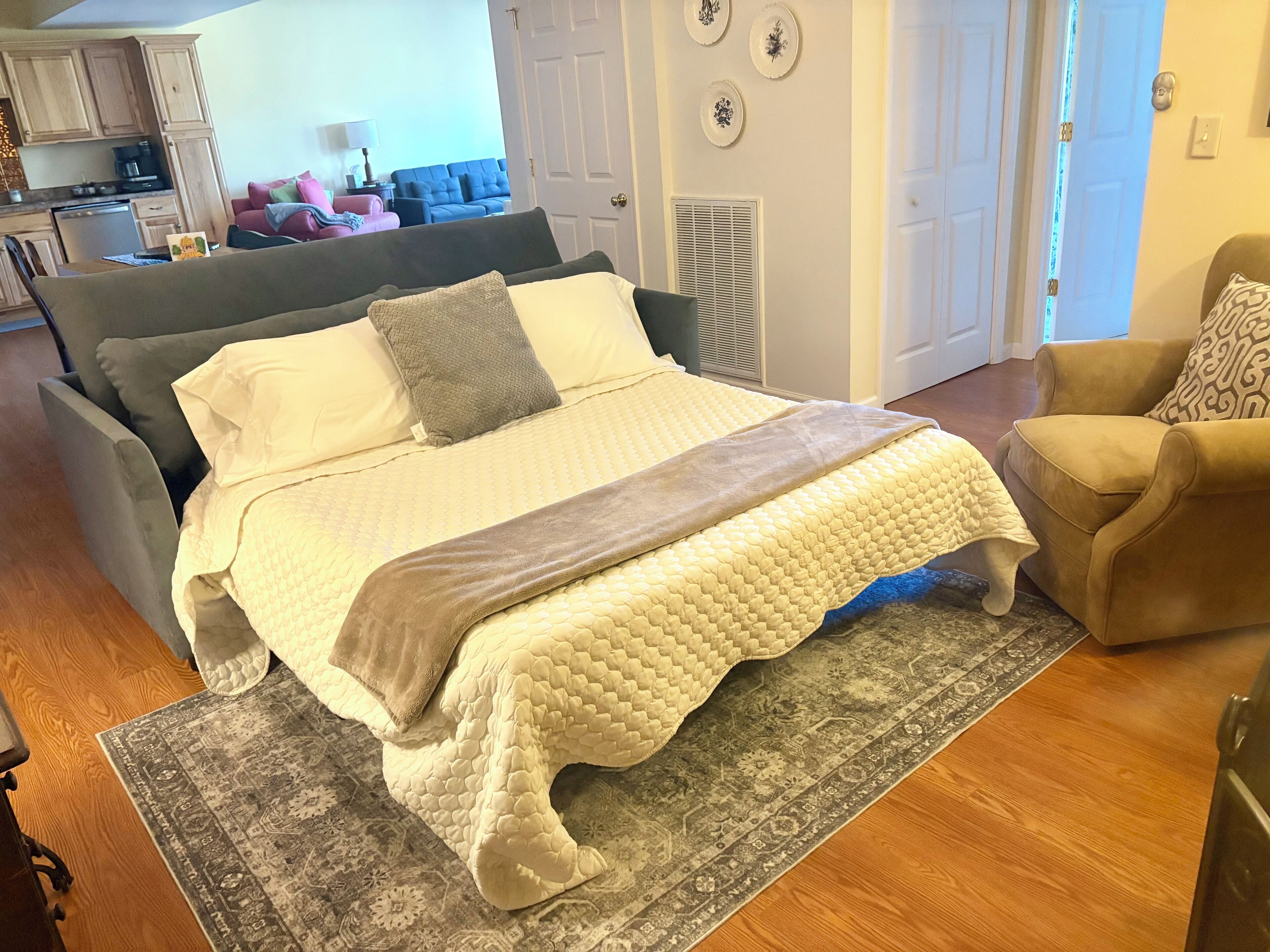 Quarto com cama, cadeira, tapete e cozinha ao fundo.