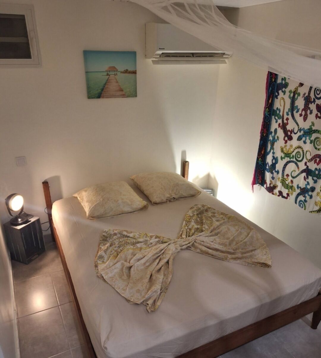 A bedroom with a bed, a nightstand, a lamp, a painting, an air conditioner, and a curtain.