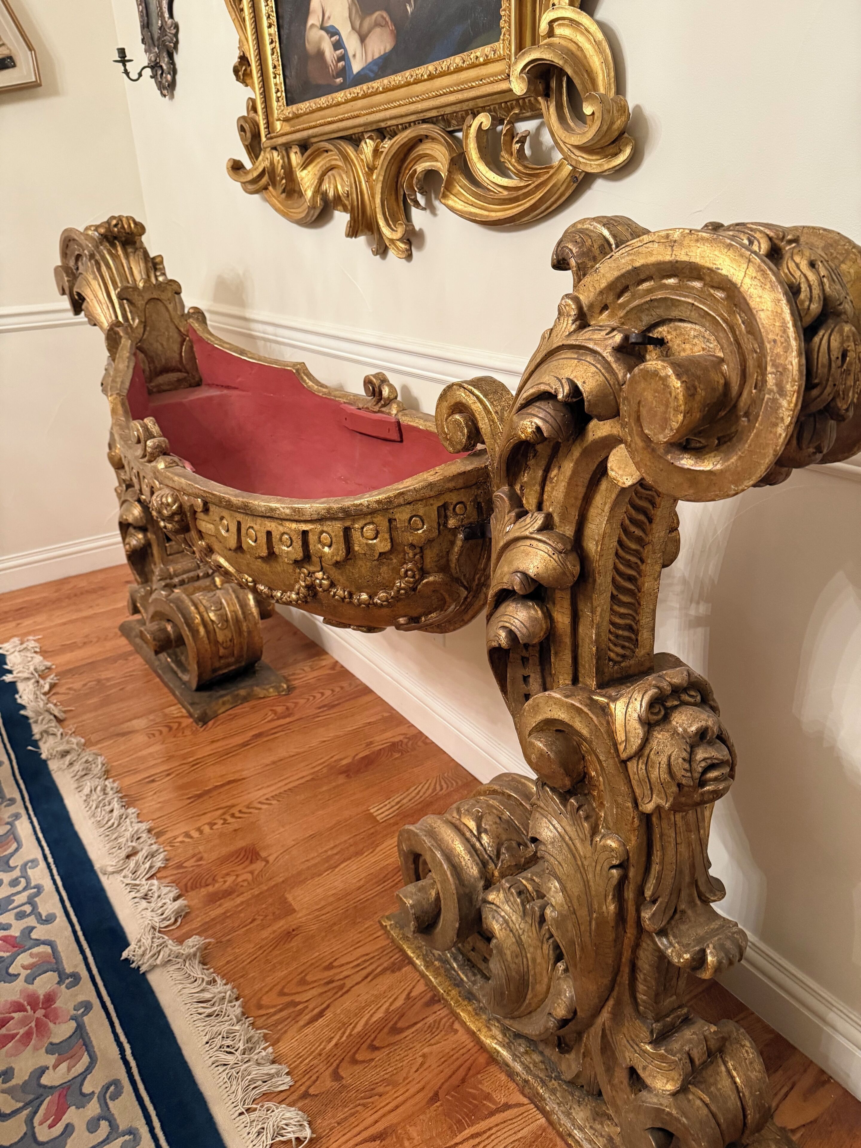 An ornate, gold-accented chair with a red cushioned seat, positioned on a wooden floor with a patterned rug.