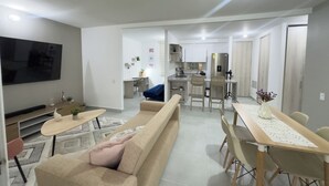 A modern living room with a sofa, coffee table, and TV. A kitchen with a dining area is visible in the background. - Mar Serena Beachside Stay  (Cartagena)