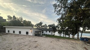 A white building with a gravel driveway and a view of a body of water. - Waterfront Elegance: Spacious 5BR Lake Home with Game Room & Private Dock Access (Osage Beach)