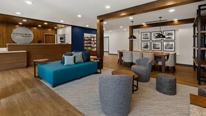 Eine moderne Hotelhalle mit einer Rezeption, einer blauen Couch, grauen Sesseln und einem Essbereich mit einem Holztisch und Stühlen.