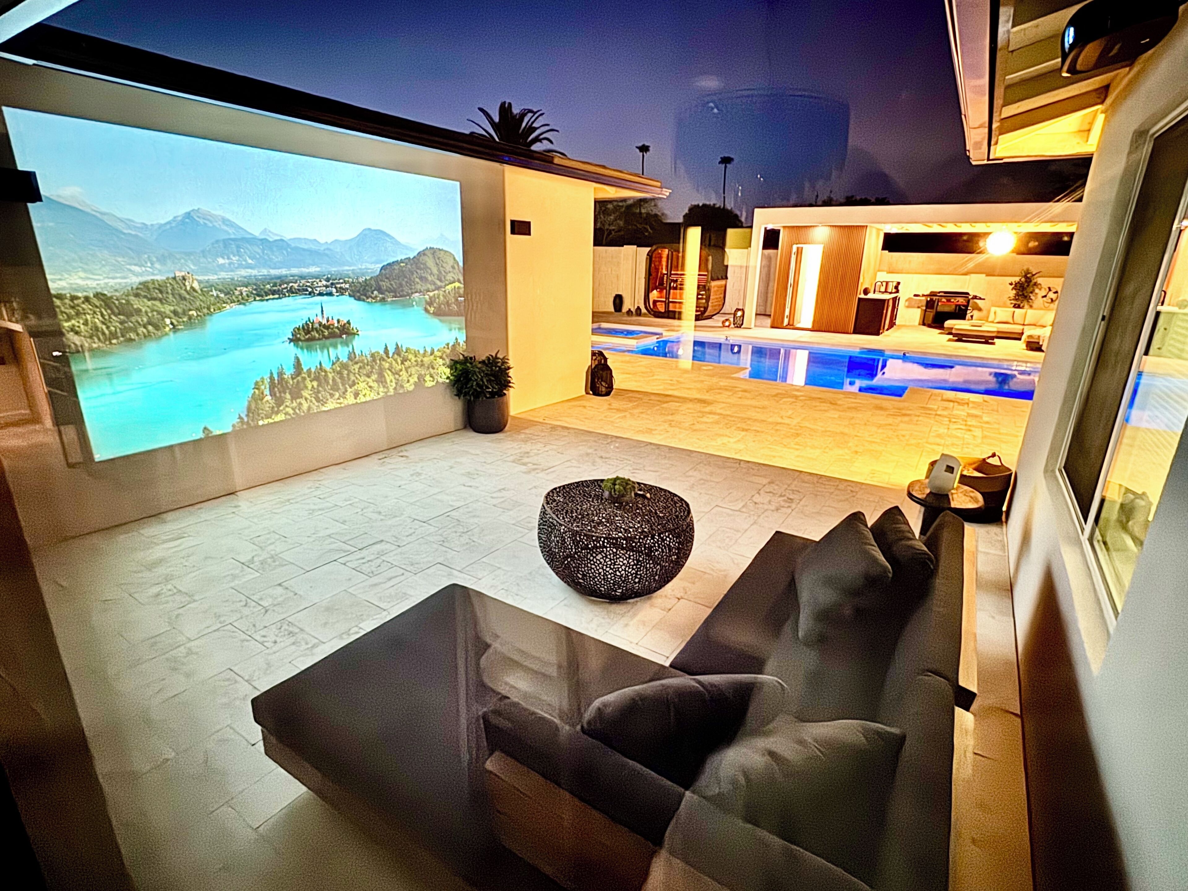 A modern living room with a large TV screen displaying a scenic landscape, a pool area with a hot tub, and a cozy seating area with a round ottoman.