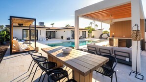 A modern outdoor patio with a pool, wooden furniture, and a seating area with cushions.