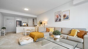 A modern living room with a sofa, coffee table, and ottoman. There is a kitchen area with cabinets and a dining table.