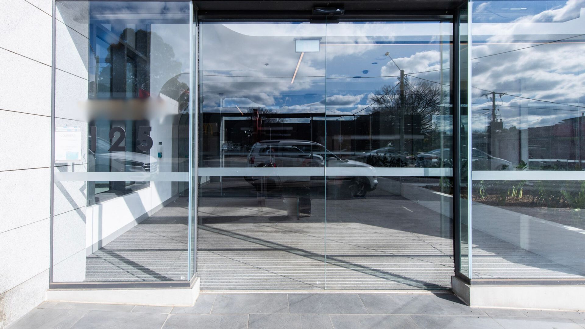 A modern building entrance with glass doors, a tiled floor, and a clear view of parked cars and a street in the background.