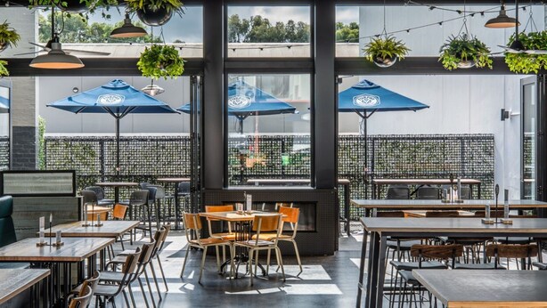 Un área de comedor al aire libre con mesas y sillas, sombrillas azules, plantas colgantes y vistas a través de amplios ventanales.