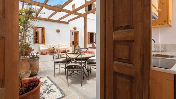 An open-plan living area with a wooden ceiling, a dining table with chairs, a sofa, and a kitchen area with wooden cabinets and a sink. - Villa Vasoula Lindos  (Lindos)