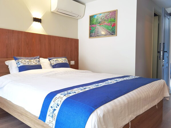 Ein Hotelzimmer mit einem Holzkopfende, einem großen Bett mit weißer und blauer Bettwäsche, einem Gartenbild an der Wand und einer Klimaanlage.