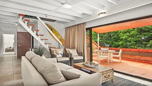 A modern living room with a staircase, a sofa, a coffee table, and a patio area with a view of greenery. - Beachfront 7 Penthouse (Sawtell)