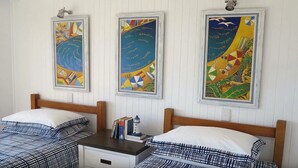 A room with two beds, each with a checkered bedspread and white pillows. There are three framed paintings on the wall, each depicting abstract maritime scenes. A small bedside table with a lamp and books is between the beds. - Beachfront 7 Penthouse (Sawtell)