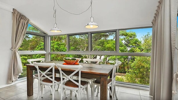 A dining area with a wooden table and white chairs, large windows with a view of greenery, and pendant lights. - Beachfront 7 Penthouse (Sawtell)