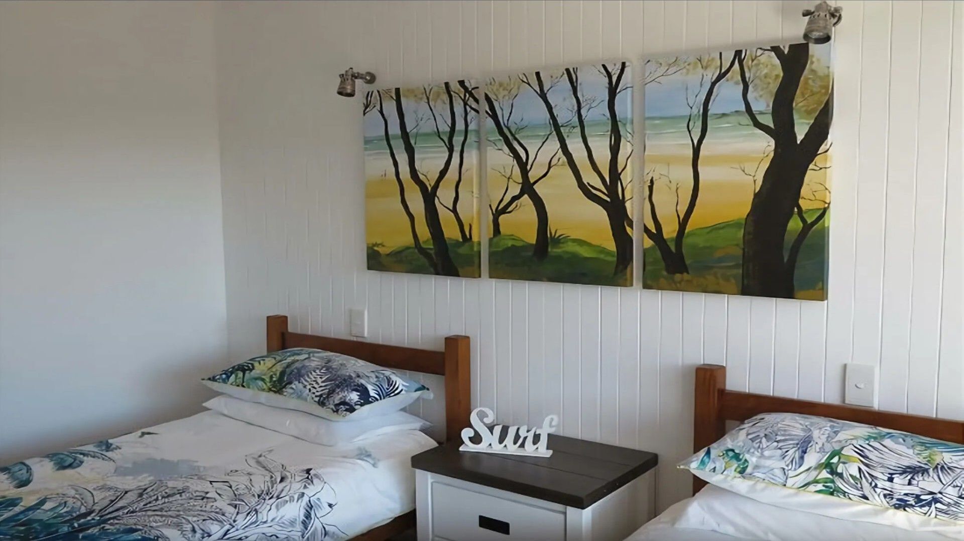 Een slaapkamer met twee eenpersoonsbedden, elk met een ander beddegoed. Aan één kant een klein nachtkastje met een lamp, aan de andere kant een decoratief bordje met de tekst 'Surf'. De muren zijn witgepanelde en boven de bedden hangt een groot schilderij van bomen en een zonsondergang.