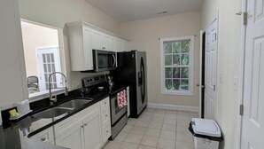 A kitchen with white cabinets, a black refrigerator, a microwave, a stove, and a sink. There is a window with a view of trees and a door on the right.