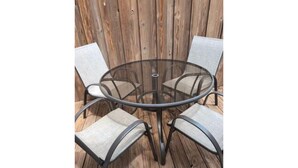 A round glass table with four chairs, each with a metal frame and fabric seat, set on a wooden deck.