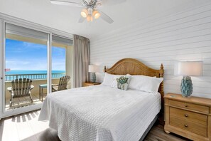 Una habitación de hotel con una cama grande, un cabecero de madera, dos mesitas de noche con lámparas, un balcón con sillas y vista al océano.