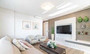 A modern living room with a sofa, two armchairs, a coffee table with flowers, a flat-screen TV, and decorative items on shelves.