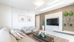Una sala de estar moderna con un sofá, dos sillones, una mesa baja con flores, un televisor de pantalla plana y objetos decorativos en estanterías.