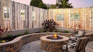 Un patio trasero con un hogar a piedra, mobiliario exterior y una valla de madera con ventanas.