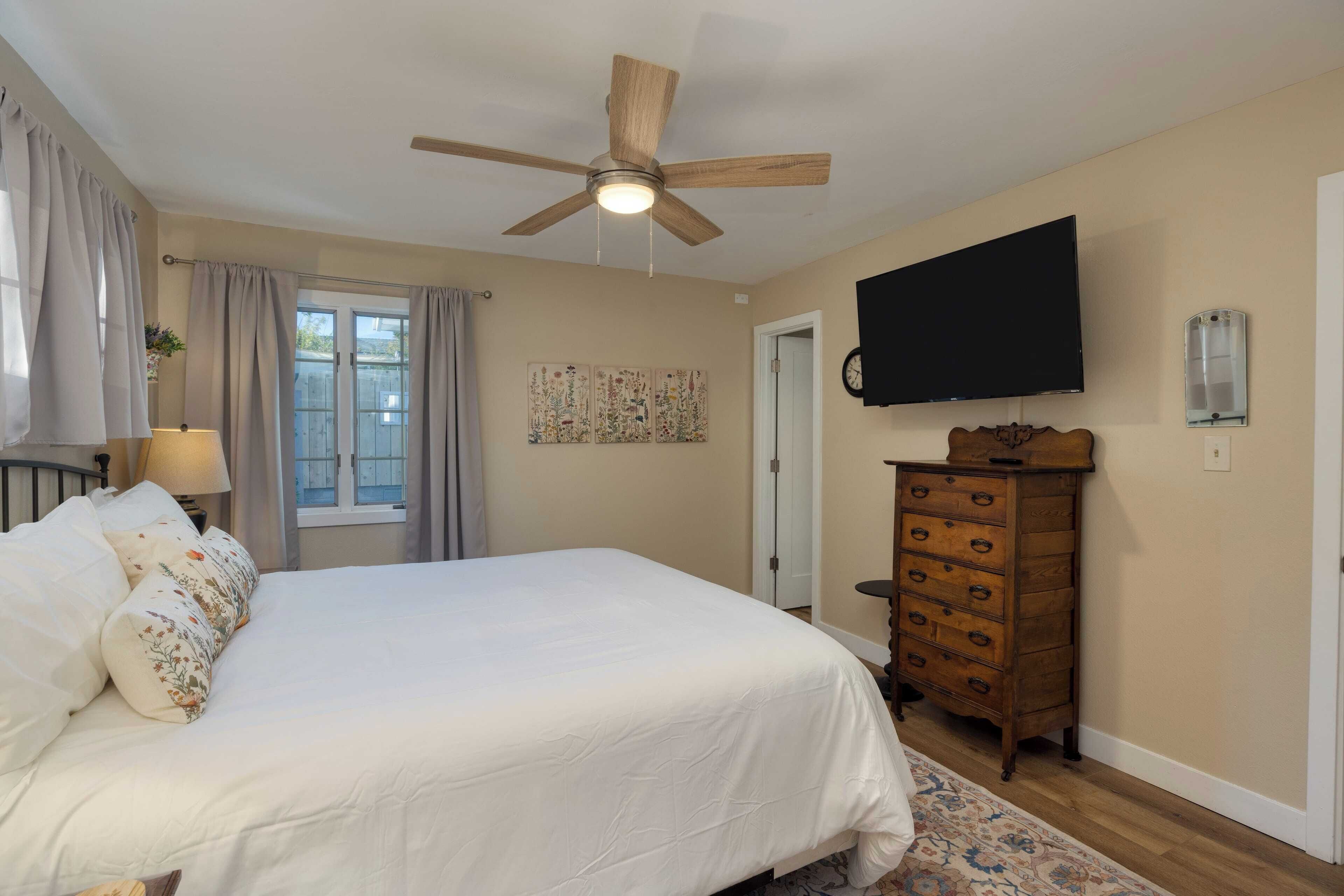 A bedroom with a bed, a ceiling fan, a dresser, a television, and a window with curtains.