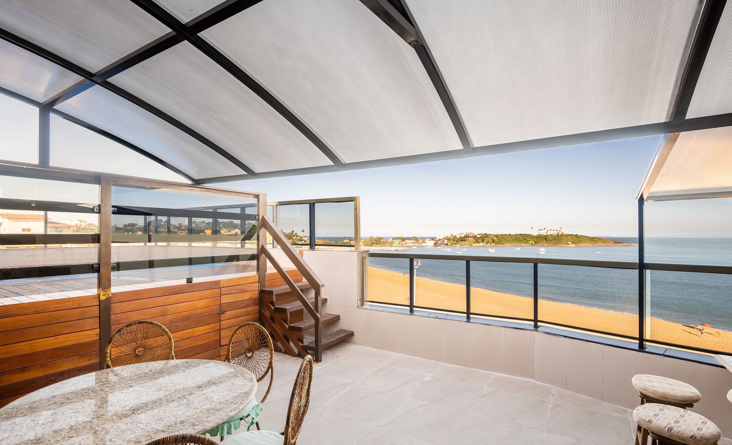 A spacious balcony with a view of the beach and ocean, wooden furniture, and a glass railing.
