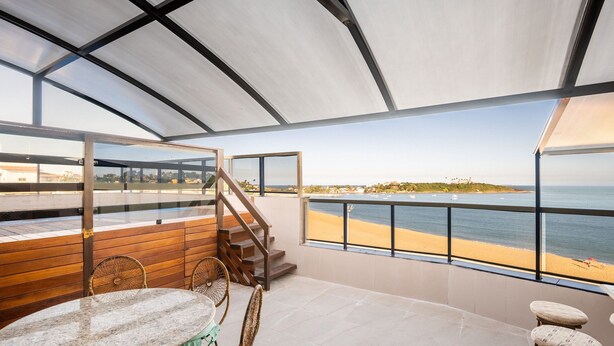 Un ampio balcone con vista sulla spiaggia e sull'oceano, arredato con mobili in legno e dotato di ringhiera in vetro.