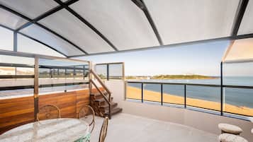 Varanda ampla com vista para a praia e o oceano, móveis de madeira e guarda-corpo de vidro.