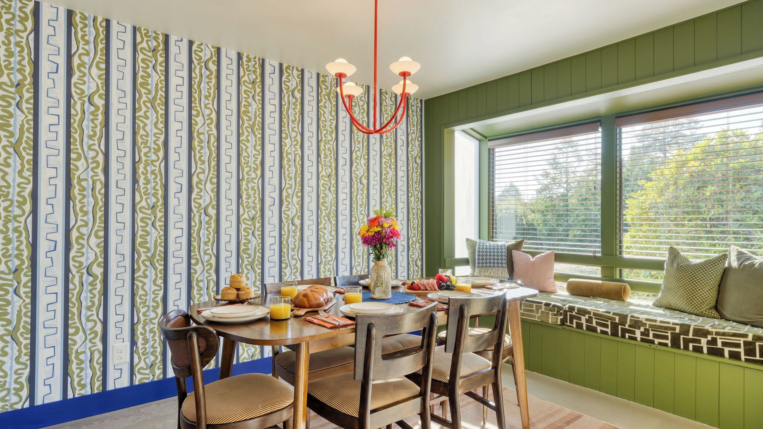 Ein Essbereich mit gemustertem Wandpapier, einer Pendelleuchte und einem Tisch, der mit Tellern, Gläsern und einer Blumenvase gedeckt ist.
