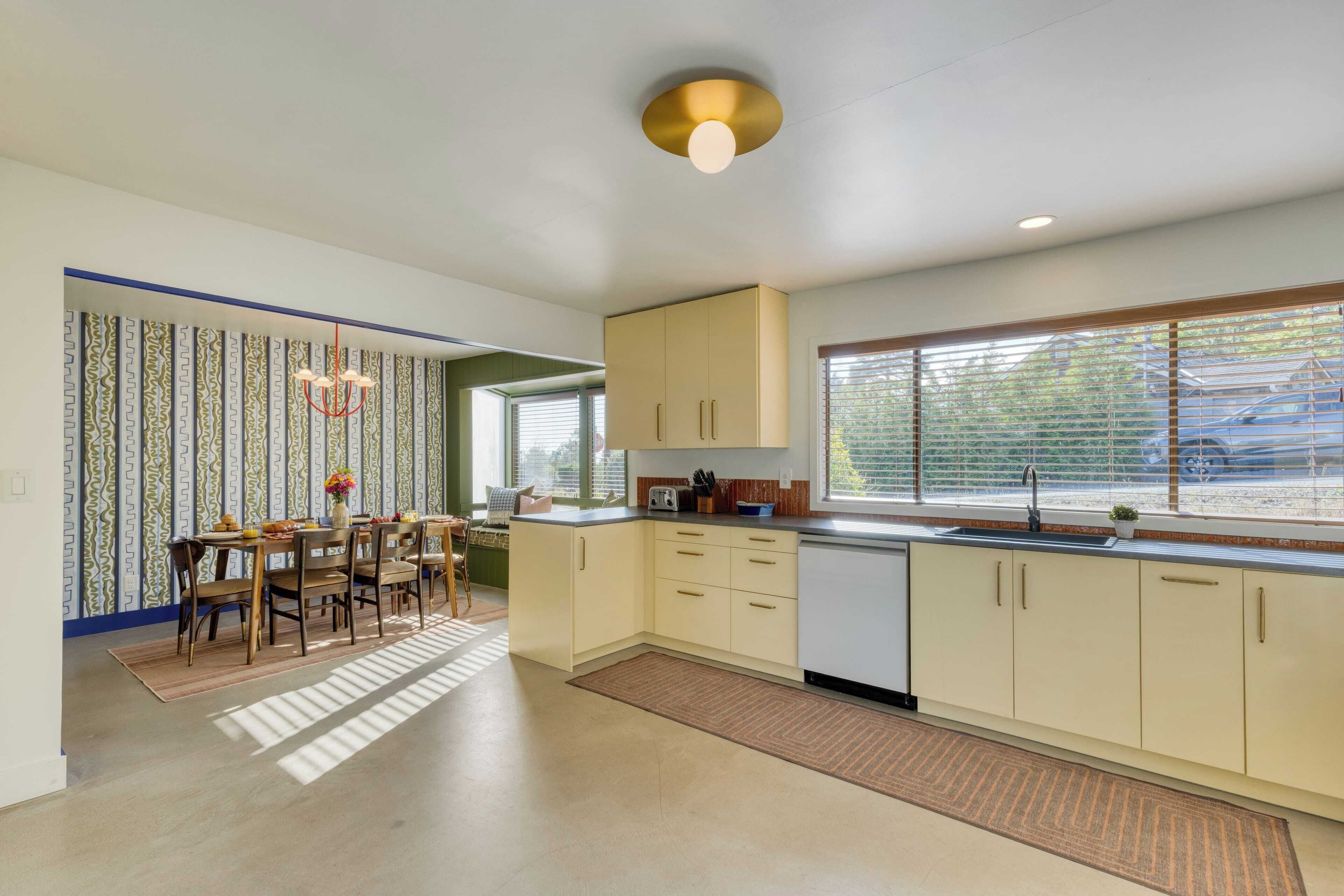 A modern kitchen with light-colored cabinets, a dining area with a table and chairs, and a window with blinds.