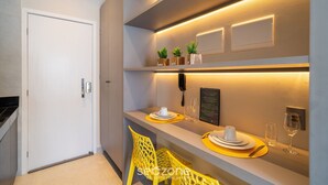 Et moderne hotelværelse med et rent, minimalistisk design. Værelset har et pænt spiseområde med gule underlayere, et hvidt bord dækket med tallerkener og bestik, samt en lille potteplante på hylden.