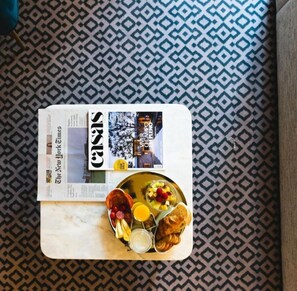A breakfast tray with croissants, fruit, and a newspaper on a patterned tablecloth.