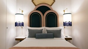 Ein Bett mit einem Kopfteil aus zwei großen, dunkel, gewölbten Feldern, flankiert von kleineren, abgerundeten Feldern. Zwei Nachttische mit Lampen sind vorhanden, jede Lampe hat einen weißen Lampenschirm und eine blaue Fransenborte. Das Bett ist mit weißer Bettwäsche bezogen, und es gibt zwei Kissen mit Muster.