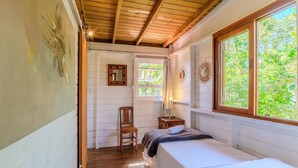 A narrow, wooden-paneled room with a bed, a chair, a small table with a lamp, and a window with a view of greenery. - House, Beach, and Nature in Ibiraquera Tan0000 (Imbituba)