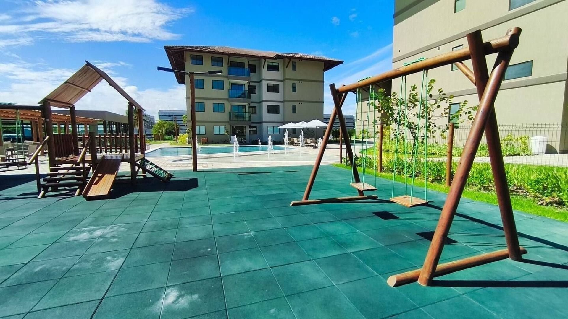 Un área de juegos al aire libre con columpios y un tobogán, una piscina y un edificio al fondo.