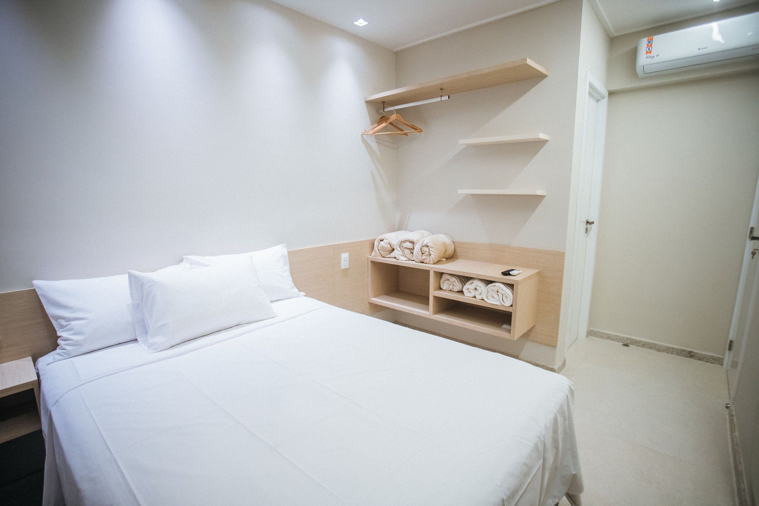 Quarto de hotel com cama, prateleira de madeira com toalhas e ar-condicionado fixado na parede.
