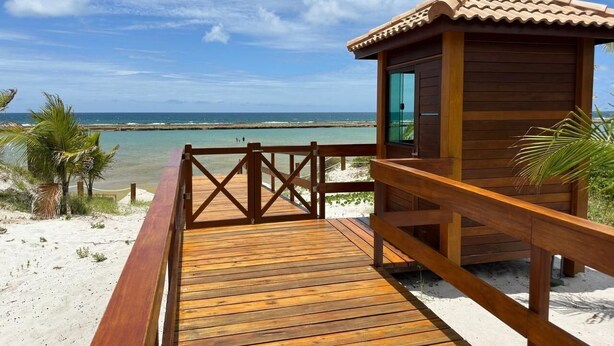 A wooden deck leading to a beachside building with a tiled roof, overlooking a sandy beach and the ocean.