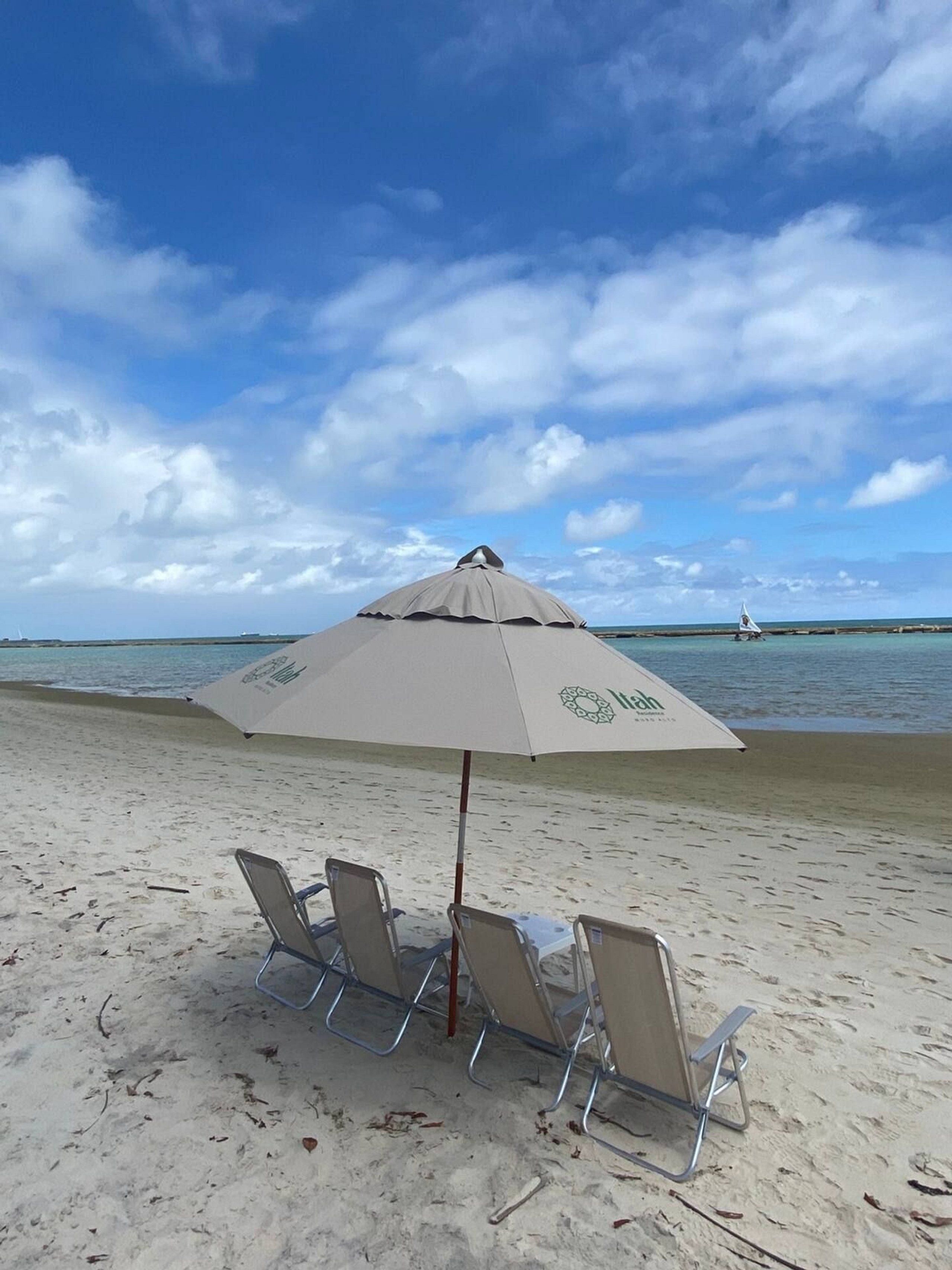Uma praia com um grande guarda-sol, três espreguiçadeiras e um veleiro ao longe.