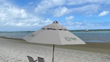 Uma praia com um grande guarda-sol, três espreguiçadeiras e um veleiro ao longe.
