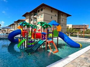 A swimming pool with a colorful play structure, including slides and climbing elements, surrounded by a building with multiple balconies. - Brisa em Muro Alto - Flat na Praia (Ipojuca)